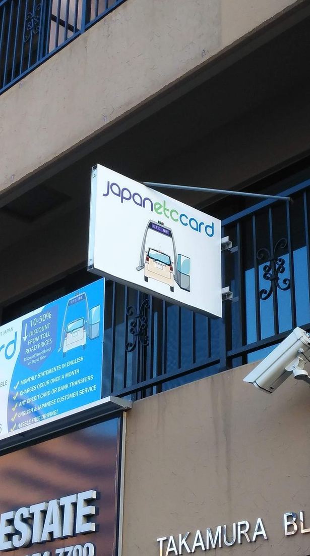 2018年新拠点のバルコニーに掲げられたJapanETCcardの吊り下げ看板 ─ シンプルなロゴと、ETCゲート通過のイラストアイコン。歩く人の目線にしっかり届く位置に。